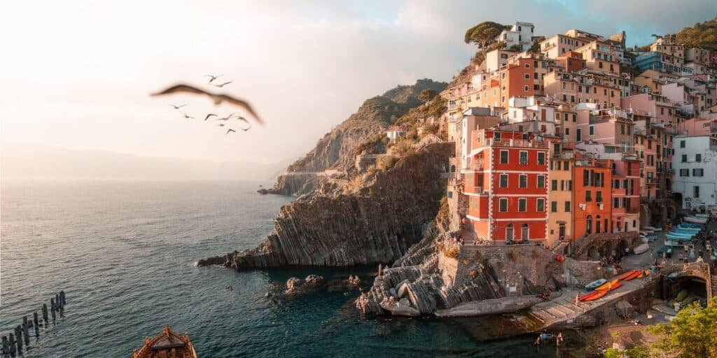 Edifícios coloridos em um litoral rochoso com vista para o mar, com pássaros voando e barcos perto da costa em Cinque Terre, La Spezia, que pode ser visitada com o eSIM Itália.