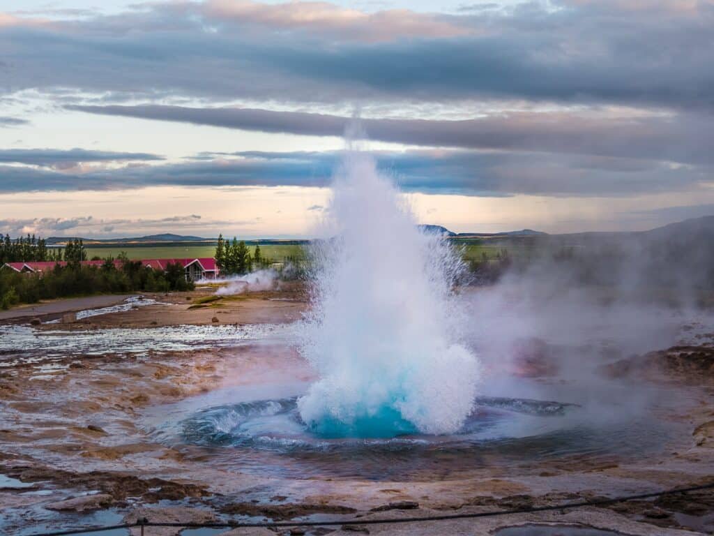 um gêiser em tons de azul rodeado por pequenas pedrinhas com construções ao fundo e um céu rasgado por nuvens com um sol leve ao fundo e alguma vegetação mais atrás, para ilustrar o post de chip celular Islândia