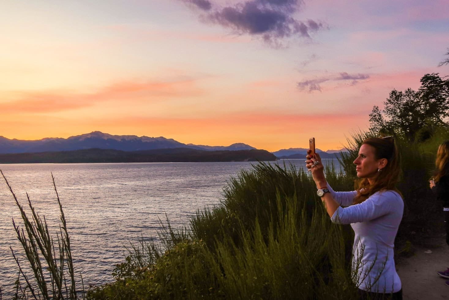 Uma pessoa tira uma foto do pôr do sol sobre um lago em Bariloche, Río Negro, Argentina, com montanhas ao fundo e vegetação ao longo da costa, que pode ser visitado com o eSIM América do Sul.