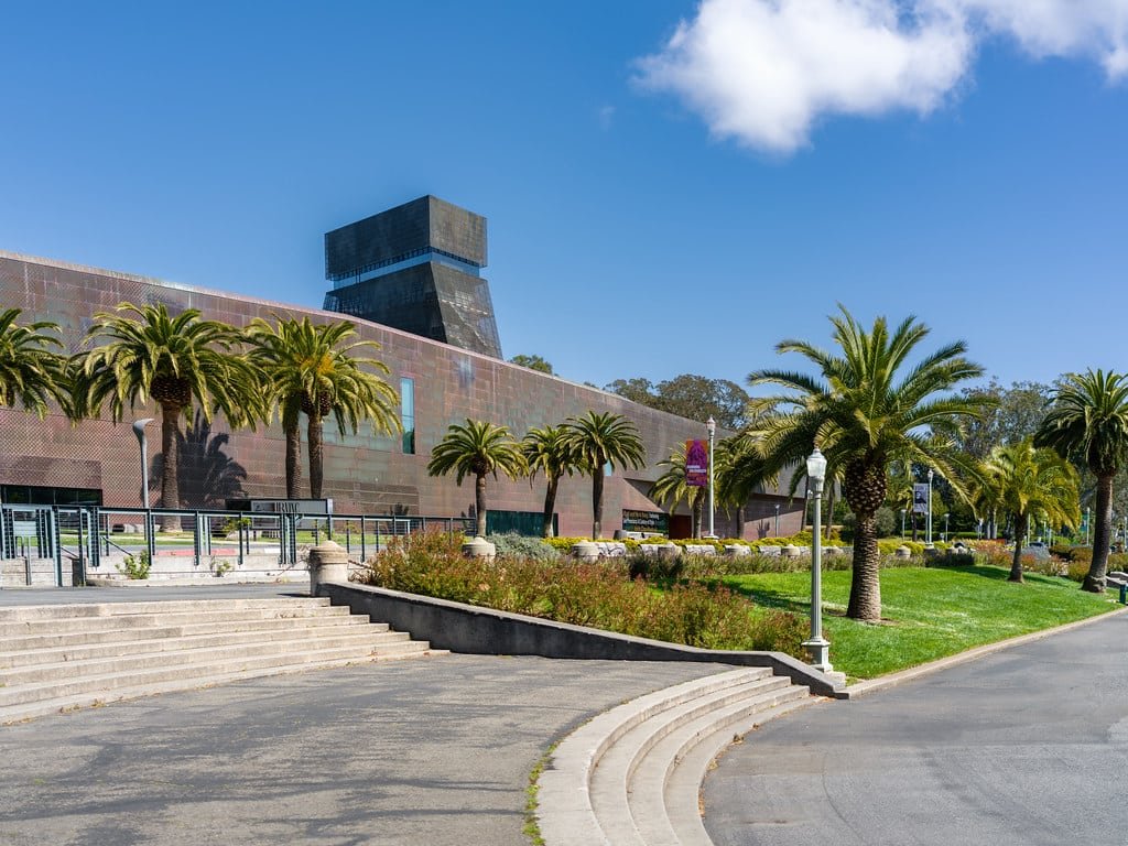 De Young Museum com fachada de cobre, ladeado por palmeiras, com um céu azul claro e cercado por jardins paisagísticos. Imagem para ilustrar post sobre museus em San Francisco.
