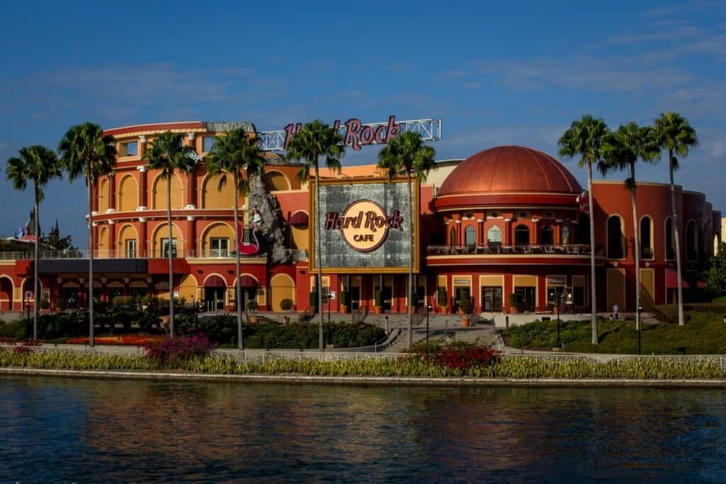 A imagem mostra a fachada do Hard Rock Cafe em Orlando, Flórida. O edifício é bastante imponente e colorido, com uma arquitetura que mistura elementos modernos e clássicos. As cores predominantes são o laranja e o vermelho, criando um visual vibrante e convidativo.

Na frente do prédio, há várias palmeiras altas, que adicionam um toque tropical ao ambiente. O logotipo do Hard Rock Cafe é claramente visível em um grande painel, que está centralizado na parte frontal da construção. Além disso, há uma grande guitarra decorativa, característica das decorações temáticas do Hard Rock Cafe, fixada na lateral do edifício.

O edifício possui várias janelas e sacadas, e o design geral apresenta um equilíbrio entre linhas retas e curvas.