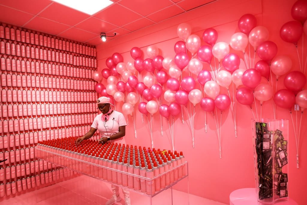 Uma pessoa está em uma sala cheia de paredes rosas, balões rosas e brancos e uma mesa coberta com latas de tinta spray vermelha do Ice Cream Museum.