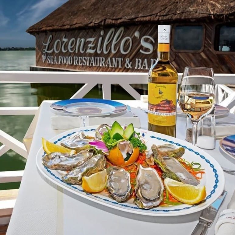 Prato de ostras com limão e uma taça ao lado de uma garrafa ao fundo. Atrás pode-se ver o restaurante Lorenzillo