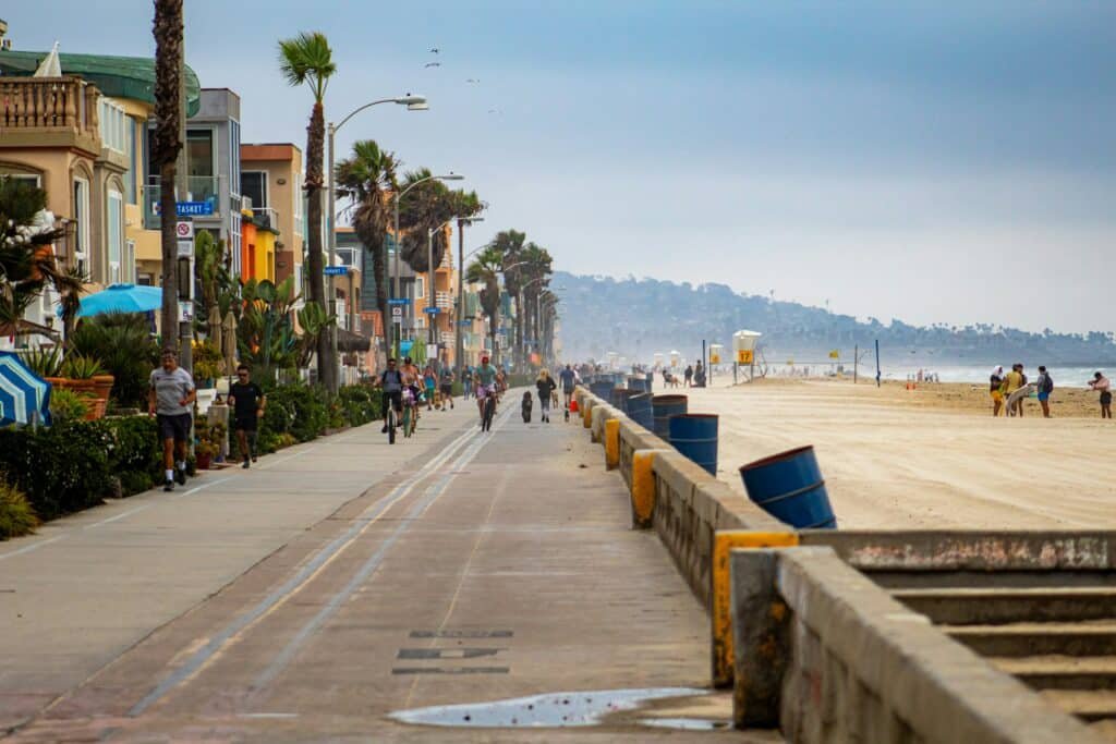 Foto da orla de Mission Beach. Várias pessoas estão andando a pé e de bicicleta pelo local, que está separado de lojas e restaurantes por alguma árvores e arbustos. Do lado direito está a praia.