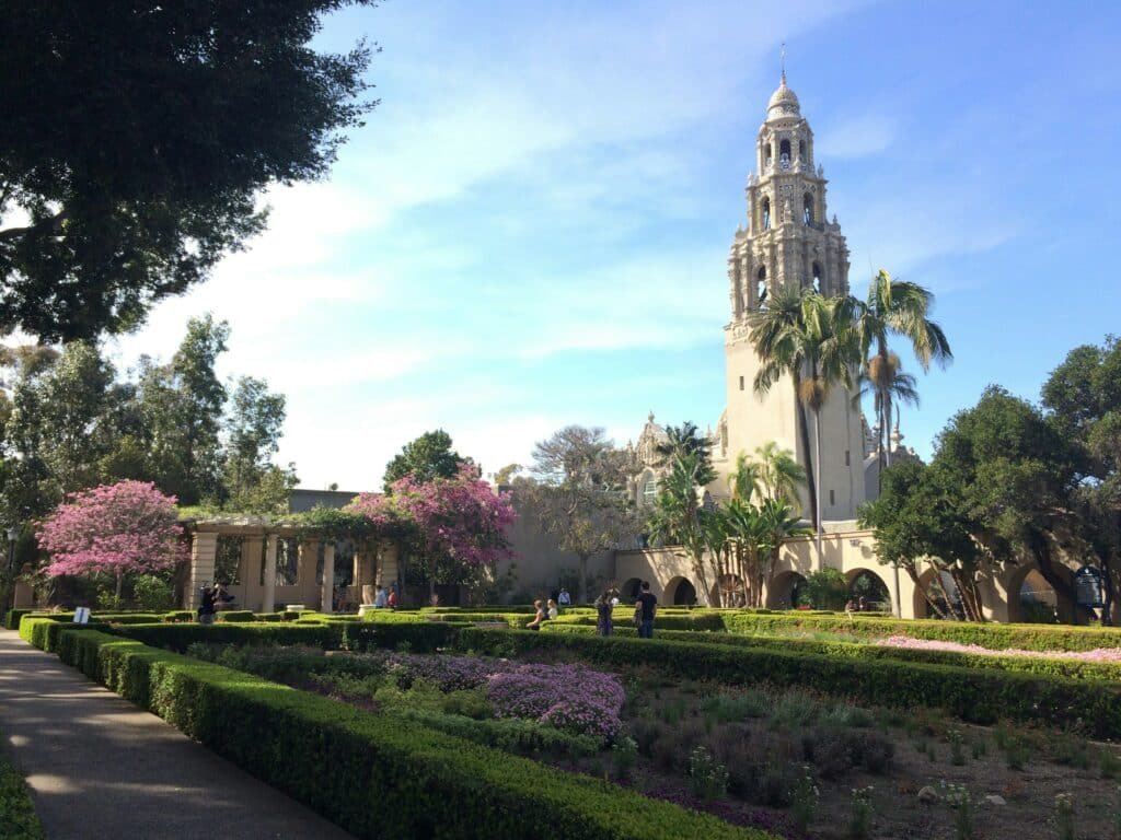 Imagem de um jardim a frente durante o dia e algumas pessoas andando e ao fundo do lado direito uma torre de uma igreja. Representa San Diego.