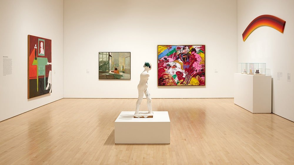 Museu de Arte Moderna de San Francisco com diversas pinturas coloridas nas paredes, uma escultura de uma figura em pé no centro e piso de madeira. Imagem para ilustrar post sobre museus em San Francisco.