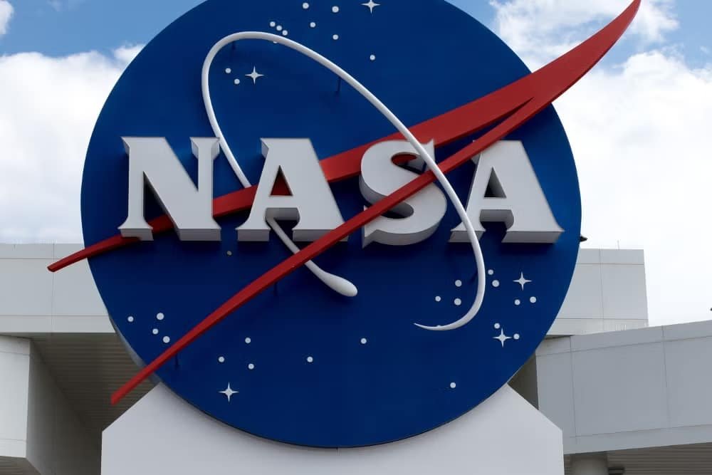 A imagem mostra o icônico logotipo da NASA no Centro Espacial Kennedy, localizado em Cabo Canaveral, Flórida. O logotipo é circular, com um fundo azul e a palavra "NASA" em letras grandes e brancas no centro. Um vetor vermelho atravessa o círculo, representando a trajetória de um foguete. Há também pequenas estrelas brancas espalhadas pelo fundo azul, simbolizando o espaço.