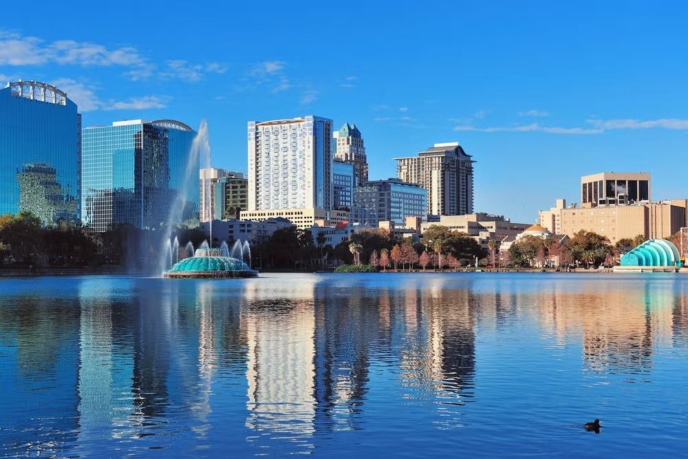 A imagem mostra uma vista panorâmica do Lago Eola, localizado no centro de Orlando, Flórida. No primeiro plano, o lago apresenta uma superfície calma, refletindo o céu azul e os edifícios ao redor. Há uma fonte grande no meio do lago, com jatos de água altos que se destacam contra o fundo.

Ao redor do lago, vários edifícios altos compõem o horizonte da cidade. Os prédios variam em estilo arquitetônico, com muitos revestidos de vidro, refletindo a luz do sol e o céu. O céu está claro, sem nuvens, indicando um dia ensolarado.