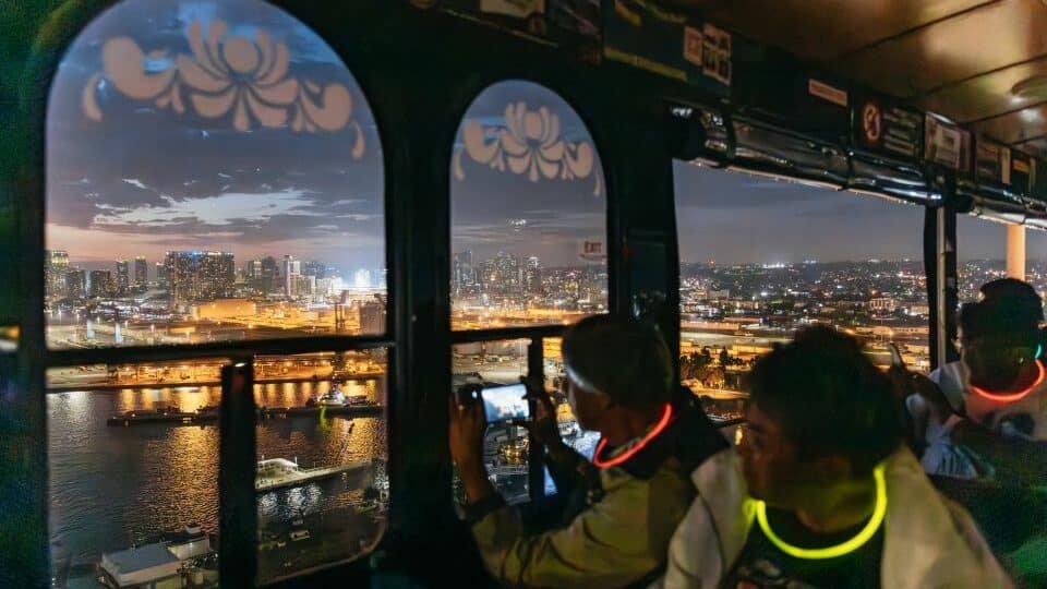 Pessoas dentro de um ônibus olhando e tirando fotos das luzes noturnas e do mar de San Diego. Todos estão com colares iluminados no pescoço. 