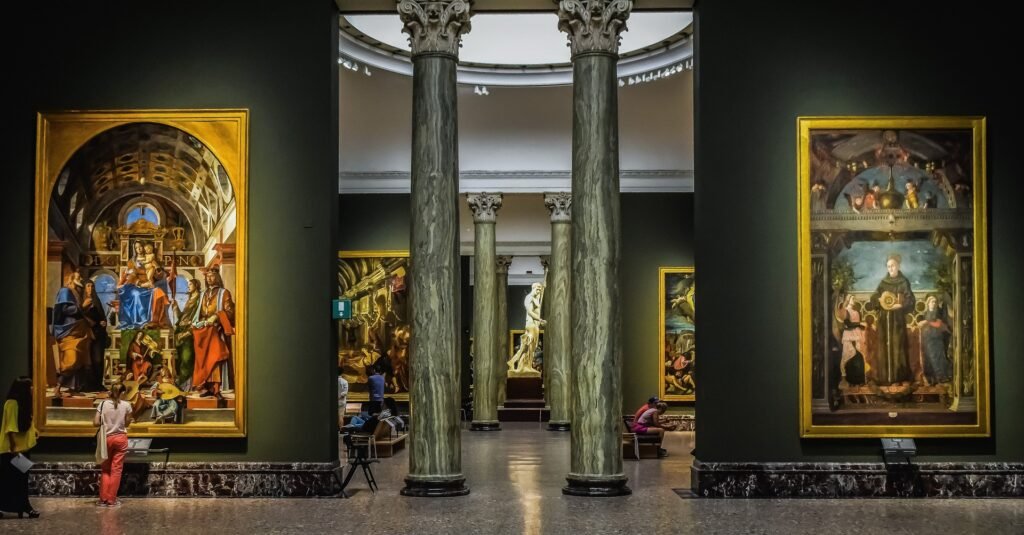 Interior de um museu de arte de Milão, a Pinacoteca de Brera, com paredes verde musgo e quadros com pinturas, colunas grossas no meio e pessoas olhando as obras de arte