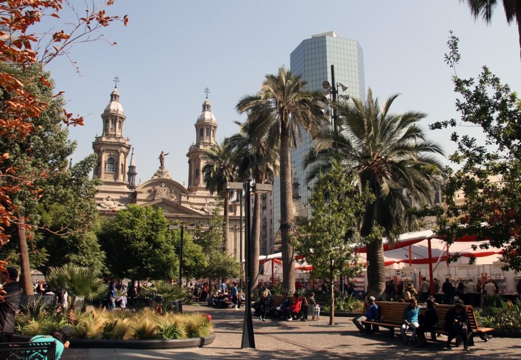 Praça em Santiago do Chile com várias árvores e plantas ao redor para ilustrar o post de seguro viagem Chile. Ao lado direito há vários bancos com pessoas sentadas conversando, e ao fundo ficam construções históricas e prédios altos. - Foto: Monica Volpin via Pixabay