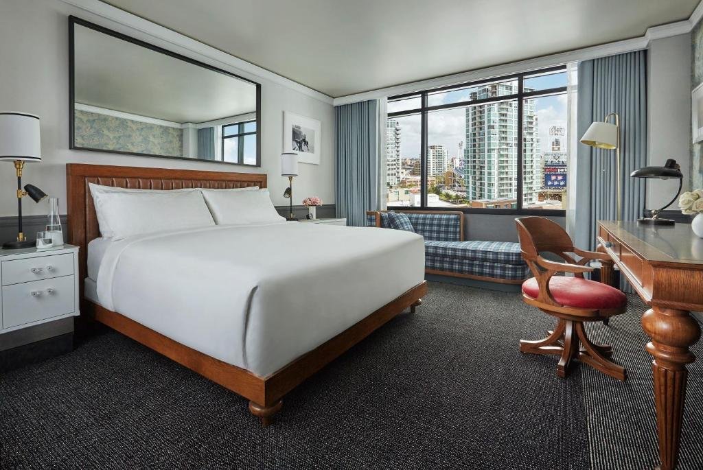 Quarto do Pendry San Diego com cama de casal ampla ao centro com espelho na parede, sofá do lado esquerdo abaixo da janela ampla de vidro com cortinas azuis, mesa de trabalho de madeira com cadeira em frente a cama e ao lado da cama duas cômodas com luminária ao lado delas. Representa San Diego.