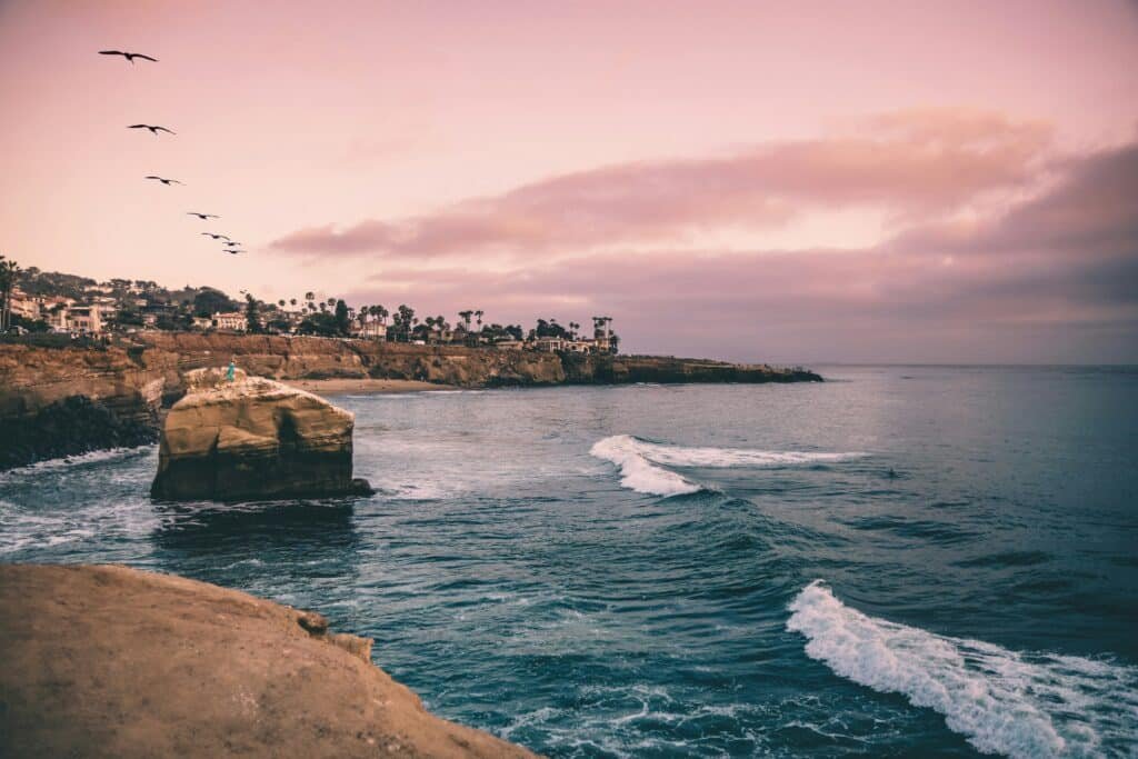 Imagem durante o dia da praia de Sunset Cliffs, com mar do lado direito do lado esquerdo rochedos e no fundo algumas árvores e casas. Representa San Diego.