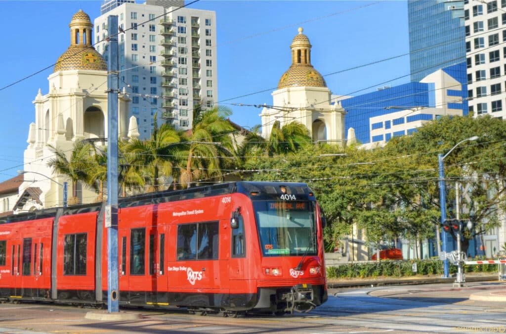 A imagem mostra o trolley andando em um trilho pelas ruas de San Diego. Ele é vermelho e igual a um trem, mas a céu aberto. Ao fundo estão algumas construções da cidade e prédios. Esse é um dos transportes em San Diego.