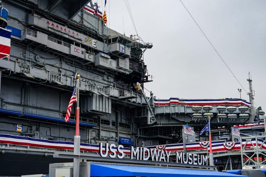 Pedaço do USS Midway Museum. Atrás do letreiro está o navio com todos os compartimentos e a bandeira dos Estados Unidos em algumas partes. A foto ilustra o post de o que fazer em San Diego.