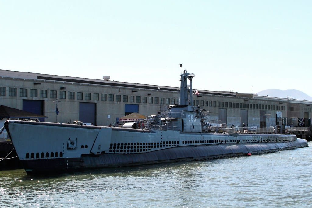 Um submarino com o número 383 na lateral está atracado perto de um grande edifício industrial perto da água.